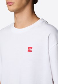 Valkoinen puuvillapaita, jossa on pieni punainen neliön muotoinen logo, jossa lukee "THE NORTH FACE". Pyöreä kaula-aukko ja lyhyet hihat.