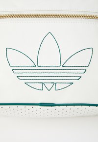 Baltas maišelis su žaliu siuvinėtu logotipu, vaizduojančiu tris lapų formos motyvus ir horizontalius bruožus, akcentuotas auksine užtrauktuku. perforuota apačia.