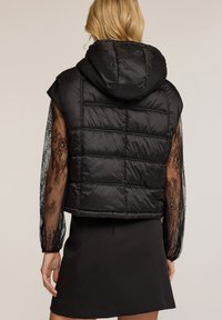 Schwarze Pufferweste mit Kapuze, kurzer Schnitt, gestepptes Design und transparenten Spitzenärmeln. Das Outfit wird durch einen schwarzen Rock ergänzt.