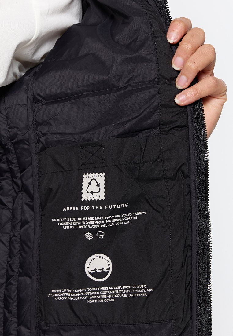 Mani che tengono aperto un giubbotto nero mostrando l'etichetta interna con il simbolo del riciclaggio e testo sui materiali riciclati e la sostenibilità oceanica.