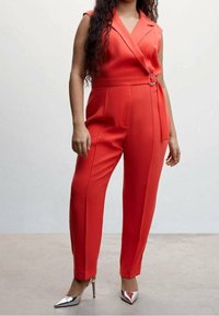 Czerwony kombinezon bez rękawów z kopertowym fasonem, bocznymi kieszeniami i paskiem. Zestawiony z metalicznie srebrnymi szpilkami na wysokim obcasie.