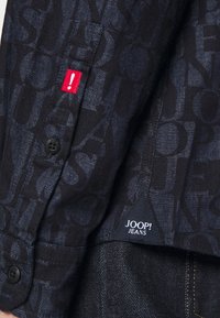 Camicia in denim nera con un motivo di lettere a contrasto, etichetta rossa in evidenza e bottone nero. L'etichetta in fondo riporta la dicitura "JOOP! JEANS."