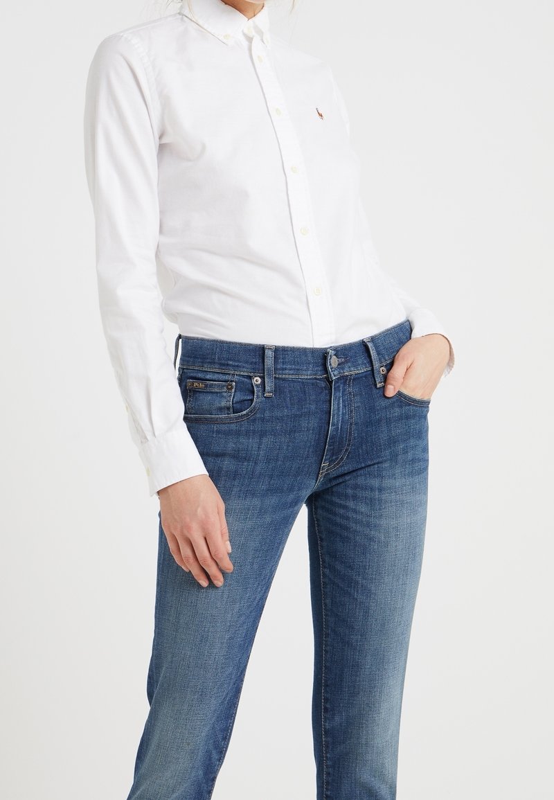 Vit skjorta med knappar gjord av bomull, som kombineras med blå denimjeans. Jeansen har en klassisk passform och subtil blekning nära knäna.