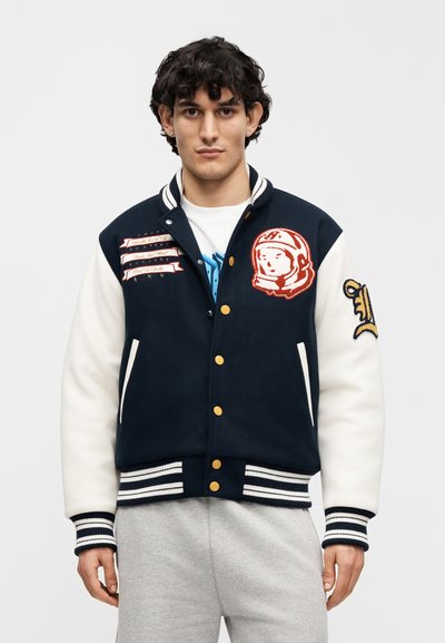 Σκούρο μπλε και λευκό varsity μπουφάν με χρυσά κουμπιά, κεντημένο αστροναύτη και ρίγες, με ριμπ μανσέτες και τελείωμα, φορεμένο πάνω από λευκό πουκάμισο.