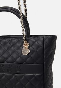 Sort quiltet tote taske med kædehåndtag, som har en gul charm og prægede logo. Glat tekstur med diamantmønster detalje.