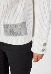 Weißer Strickpullover mit geripptem Muster, versehen mit einem silbernen Grafikpatch und zwei dekorativen Knöpfen am Ärmel. Kombiniert mit schwarzen Hosen.