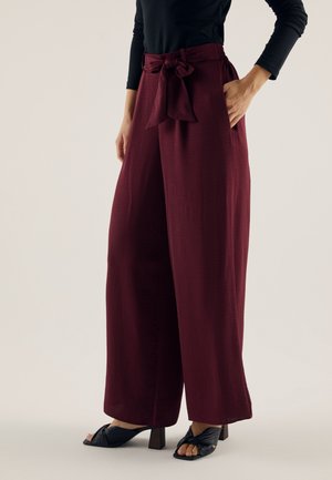 Femme portant un pantalon bordeaux taille haute à jambes larges, avec une ceinture en tissu nouée en nœud papillon, associée à des sandales noires à talons et un haut noir.