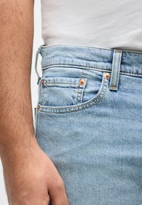 Κοντινό πλάνο ενός ατόμου που φοράει τζιν παντελόνι ανοιχτού μπλε χρώματος με χάλκινα καρφιά και λευκό πουκάμισο μπουρντουρισμένο, δείχνοντας το αριστερό του χέρι δίπλα του.