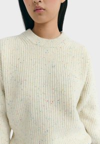 Frau trägt einen cremefarbenen Strickpullover mit einem gerippten Muster und mehrfarbigen Sprenkeln vor einem schlichten weißen Hintergrund.