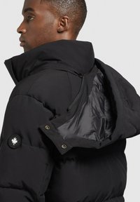 Schwarze Daunenjacke mit hohem Kragen, glänzendem Innenfutter, Reißverschluss und runder Logopatch auf der linken Schulter.