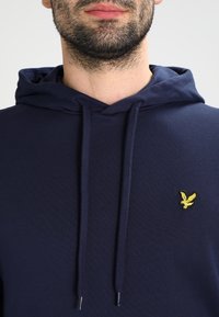 Hoodie bleu marine avec une poche avant et un cordon de serrage. Dispose d'un petit logo brodé jaune sur la poitrine gauche. Texture de tissu douce.