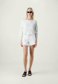 Sweater curto branco com detalhes texturizados, combinado com calções de denim brancos desgastados. Modelo a usar óculos de sol pretos e sandálias tipo slide.