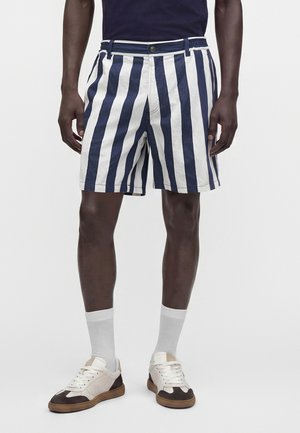 Bărbat purtând pantaloni scurți cu dungi verticale bleumarin și albe, șosete albe, adidași albi cu tălpi maro și o cămașă închisă la culoare, stă în picioare, drept.