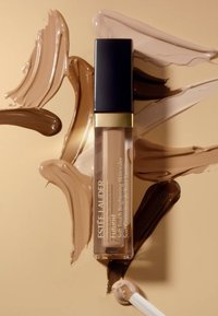 Estée Lauder Futurist concealer i en klar flaska med en guldaccent, omgiven av olika krämfärger på en beige bakgrund.