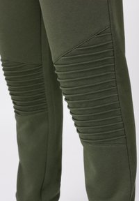 Pantaloni sport kaki-verzi confecționați dintr-un material moale, cu panouri côtelées pe coapse, având un design elegant conic și o textură netedă.