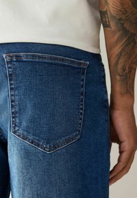 Blugi din denim albastru închis cu un buzunar la spate. Cusutura este de un maron distinctiv. Materialul pare neted, cu o ușoară textură.