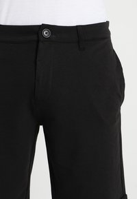 Pantalones negros con cierre de botones en la parte delantera, dos bolsillos laterales y una textura suave. La tela parece ligera y versátil para el uso casual.