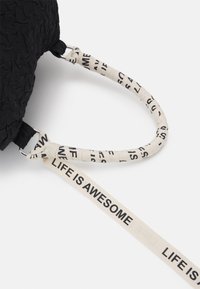 Zwart gestructureerde tas met een beige riem. Op de riem staat de tekst "LIFE IS AWESOME" in het zwart gedrukt. Metalen accenten verbinden de riemen.
