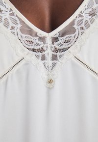 Λευκό πουκάμισο με V-σχήματος λαιμόκοψη, διακοσμημένη με περίπλοκες δαντελένιες λεπτομέρειες, μικρό διακοσμητικό κουμπί και διακριτές ραφές κατά μήκος των ραφών.