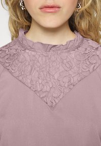 Lavendelblus med en texturerad spetspanel i V-form vid bröstet, med en volangkrage och slät tygfinish.