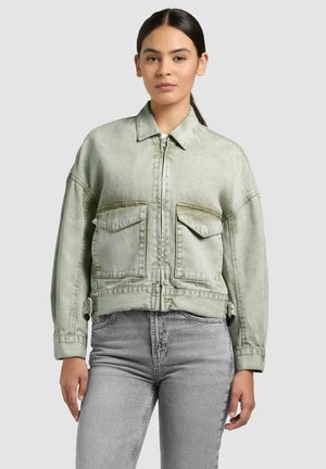 Fiatal nő világoszöld cipzáras dzsekiben nagy első zsebekkel és világosszürke farmernadrágban, egyszínű szürke háttér előtt áll.