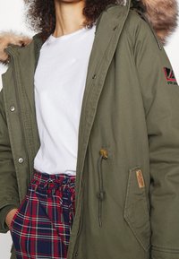 Parka verde oliva con cappuccio in pelliccia sintetica, zip frontale e chiusura a scatto. Presenta una vita regolabile con cordino e pantaloni a quadri con motivi rossi e blu navy.