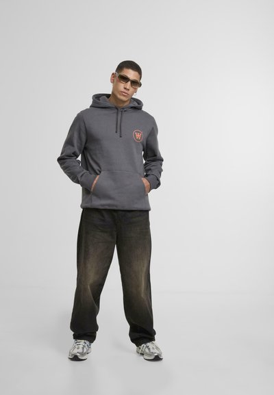Grå hettegenser med justerbar snor, frontlomme og oransje logo, kombinert med løse sorte jeans og grå sneakers. Jevn tekstur.
