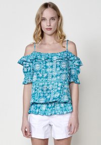 Top verde acqua con spalle scoperte, maniche a balza e design a fantasia. Realizzato in tessuto leggero, abbinato a pantaloni corti bianchi.