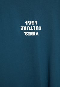 Tyrkysová látka s bílým nápisem „VIBES. CULTURE 1661“ tištěným vzhůru nohama.