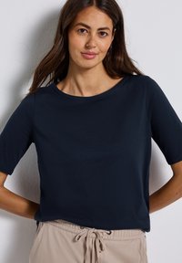 Femme aux longs cheveux bruns portant un haut à manches courtes bleu marine et un pantalon beige avec cordons, se tenant contre un mur blanc.