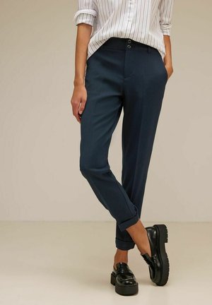 Frau trägt marineblaue, maßgeschneiderte Hosen mit aufgekrempelten Säumen, ein in die Hose gestecktes weißes gestreiftes Hemd und schwarze klobige Loafers, steht mit einer Hand in der Tasche.