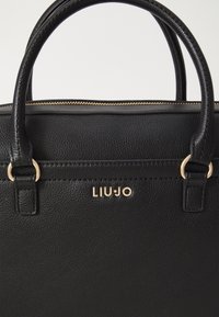 Sac à main en cuir noir avec surface texturée, accessoires en métal doré et double poignées supérieures. Présente le nom de la marque "LIU JO" sur le devant.