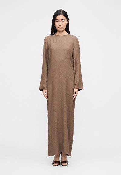 Γυναίκα με μακρύ σκούρο μαλλί, που φοράει καφέ φόρεμα μακρυμάνικο μέχρι το πάτωμα και καφέ πέδιλα με ανοιχτά δάχτυλα, στέκεται μπροστά σε λευκό απλό φόντο.