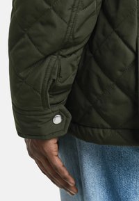 Dunkelgrüner gesteppter Jackenstoff mit einer Nahaufnahme eines Ärmels, der einen silbernen Druckknopf zeigt, und einer Hand, die auf dem Rand einer Jeanshose ruht.