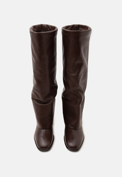 Paire de bottes hautes en cuir marron foncé avec des bouts ronds, debout sur un fond clair uni.