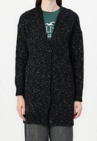 Svart stickad cardigan med en strukturerad design, ribbade muddarna och ett knappstängning, med kontrasterande vita fläckar genom hela.