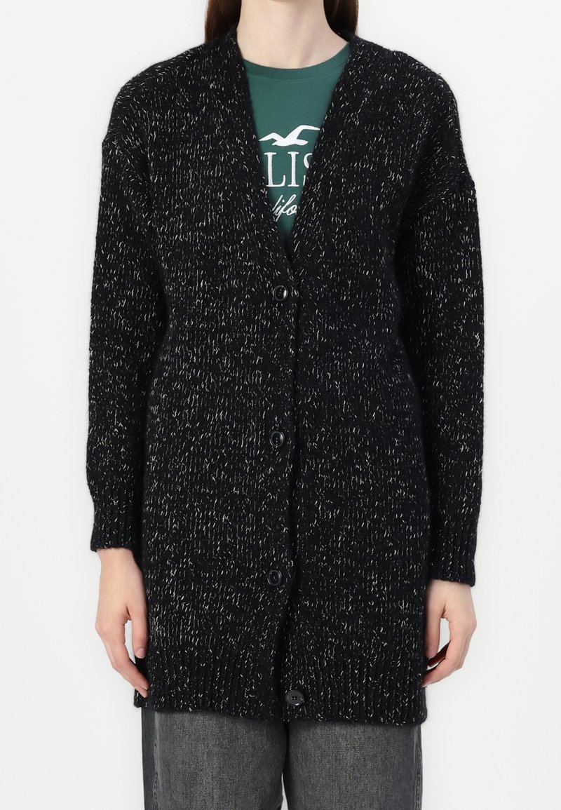 Svart stickad cardigan med en strukturerad design, ribbade muddarna och ett knappstängning, med kontrasterande vita fläckar genom hela.