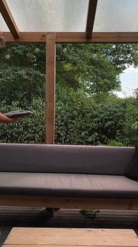 Mörkgrå vadderad soffa med träram, som har ett texturerat kastkudd. Utomhusmiljö med grönska i bakgrunden och trägolv.