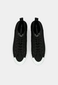 Μαύρα ψηλά αθλητικά παπούτσια με υφασμάτινο επάνω μέρος, λευκή λαστιχένια μύτη και λευκή σόλα. Διαθέτουν μαύρα κορδόνια και λογότυπο στη γλώσσα του παπουτσιού.