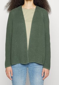 Knit cardigan in dark green, open front, textured fabric with vertical ribbing and a loose fit. Paired with a light green shirt and jeans.