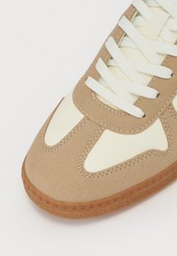 Tan ruskinds sneaker med creme lærredsfunktioner, rund tå, flade hvide snørebånd og tekstureret gummisål med mønsterkant.