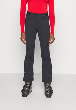 Personne portant un pantalon de ski noir avec des genoux rembourrés, des bottes de ski noires et un haut à manches longues rouge, debout devant un fond uni.