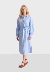 Vestito camicia blu a righe con vita alta da annodare, bottoni frontali, maniche lunghe e spacchi laterali. Indossato con sandali marroni con cinturini.