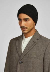 Zwarte geribbelde gebreide muts gecombineerd met een bruine getextureerde blazer met een inkeping revers en een enkele knoopsluiting. Witte henley shirt zichtbaar.