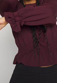 Bordeauxfarbene Bluse mit langen, gepufften Ärmeln, gerafften Manschettendetails und einer gerafften Taille, aus leichtem Stoff. Getragen mit schwarzen Hosen.