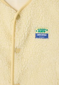 Kreminės spalvos fleece liemenė su minkšta tekstūra, apvaliu apykakliu ir kreminiais mygtukais. Turi mėlyno ir žalio „Super Vintage“ etiketę.