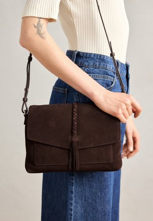 Kvinde iført blå jeans og cremefarvet ribbet top, der holder en mørkebrun ruskind crossbody taske med fletdetalje på forsiden.