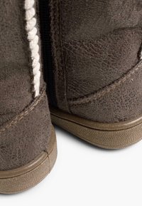 Brązowe zamszowe botki za kostkę z plecionym białym detalem, suwakiem po boku i teksturowaną gumową podeszwą. Widoczne szwy i zużyta faktura powierzchni.