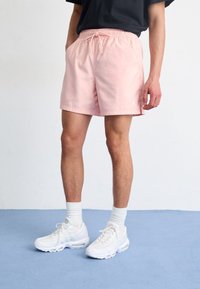 Man i ljusrosa shorts, vita sneakers och vita strumpor som står på ett blått golv med händerna i fickorna mot en vit vägg.
