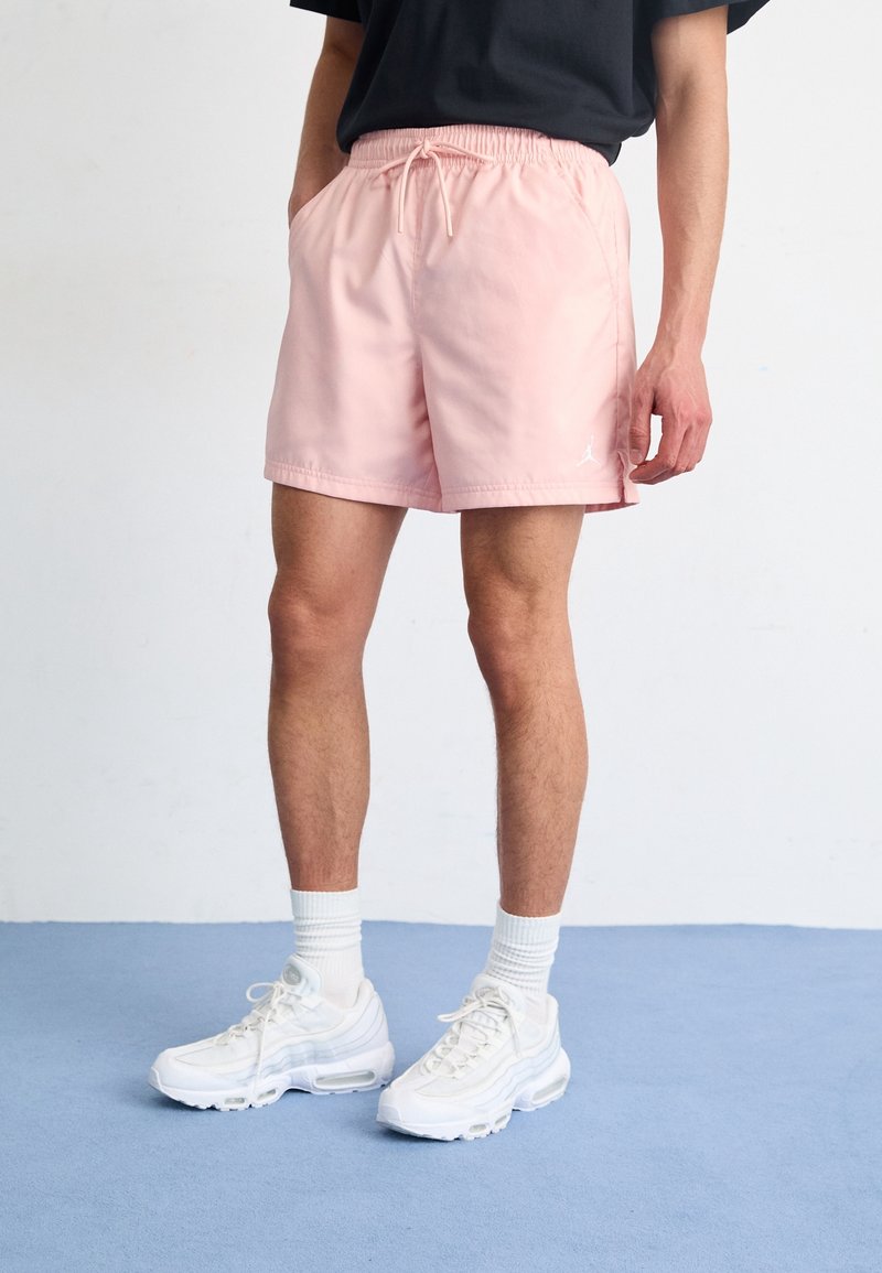 Man i ljusrosa shorts, vita sneakers och vita strumpor som står på ett blått golv med händerna i fickorna mot en vit vägg.